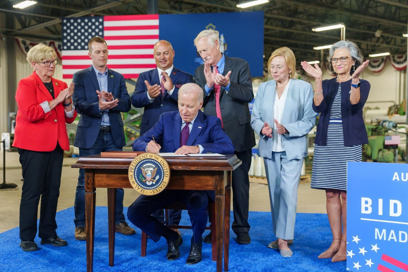 President Biden, seated and signing a document surrounded by individuals in his visit to Auburn Manufacturing in Auburn, Maine.